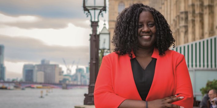 A photograph of a Black woman stood next to the River Thames and the Palace of Westminster. She is wearing a black shirt and a red jacket with her arms folded. She is smiling.