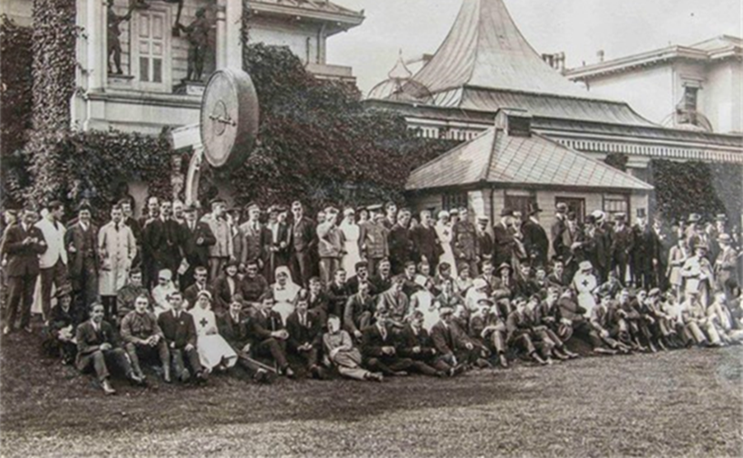 A black and white photograph of a large group of people (80-100 people). Some of the group are women wearing nurses uniform, and some are men wearing military uniform, and men wearing suits. Half are stood and half sat on grass (St Regent's Park) in front of a large building.
