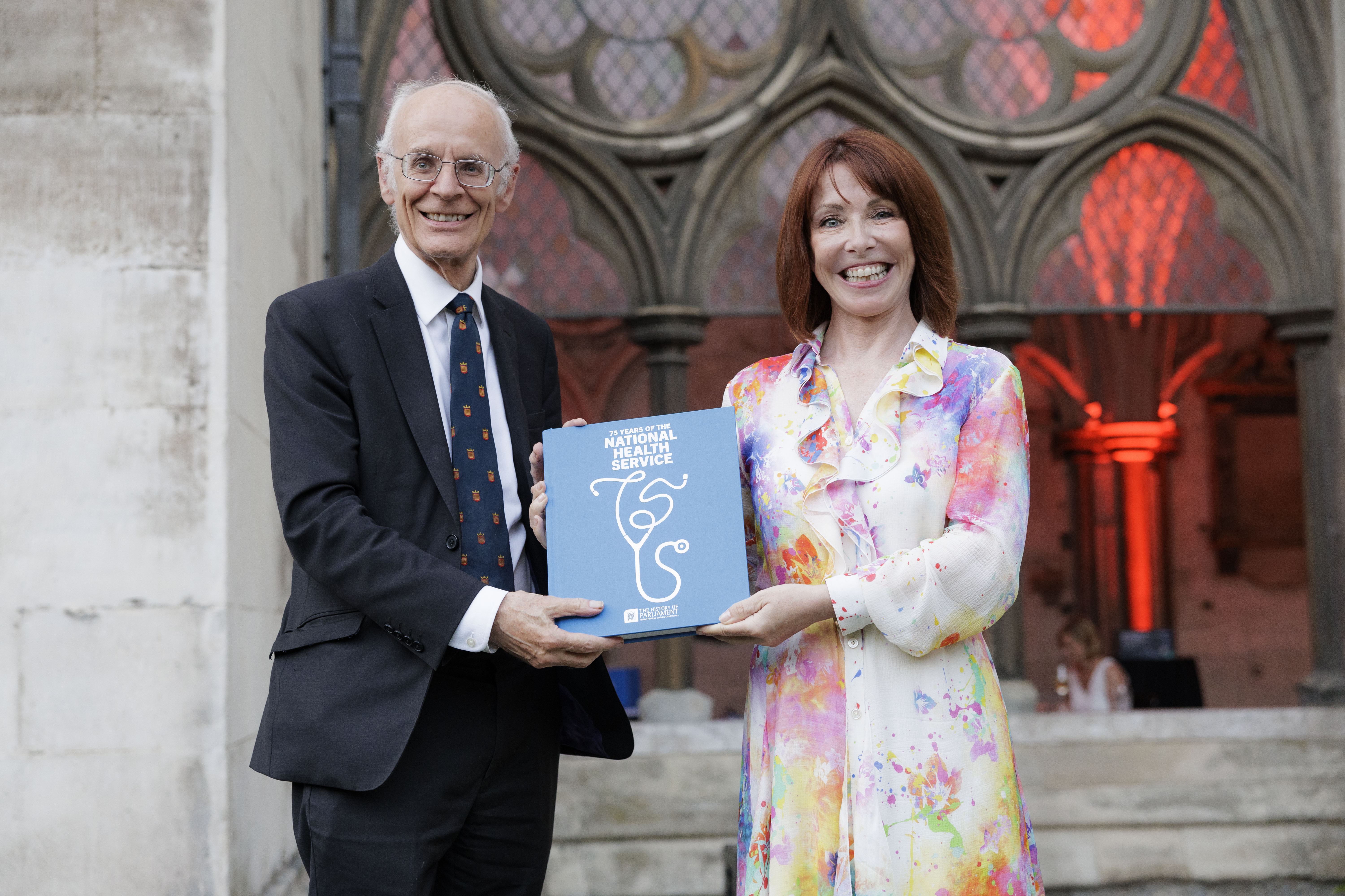 A man in a navy suit and navy tie and a woman with red hair and a multicoloured dress stand smiling, facing the camera, with a blue book between them. An elaborate arched window is in the background.