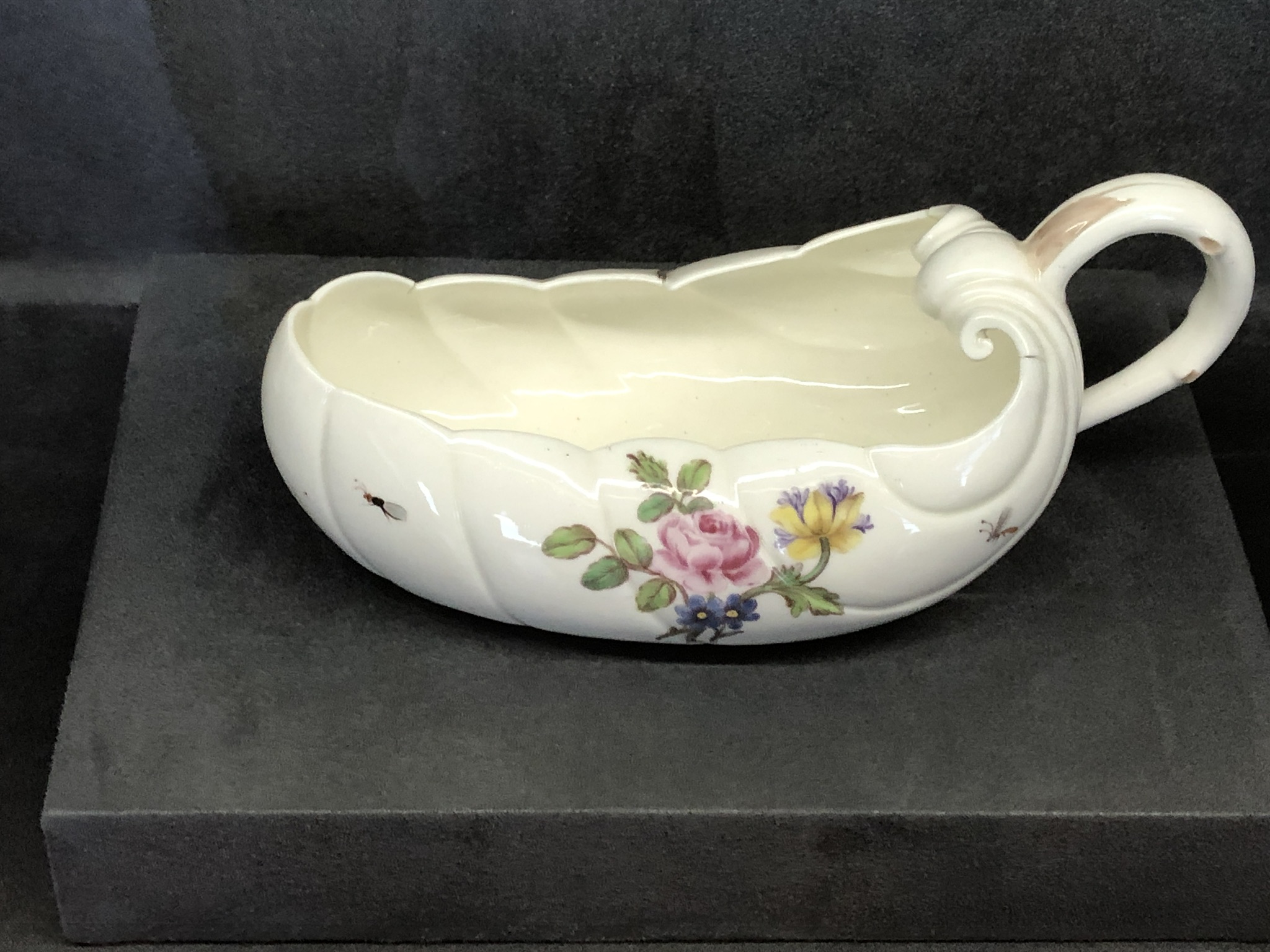 A photograph of a bourdaloue pot. It is a boat-shaped vessel with a raised lip at one end and handle at the other, a bit like a gravy boat. It is white and there is a painting of flowers on the front, with bees flying around it.