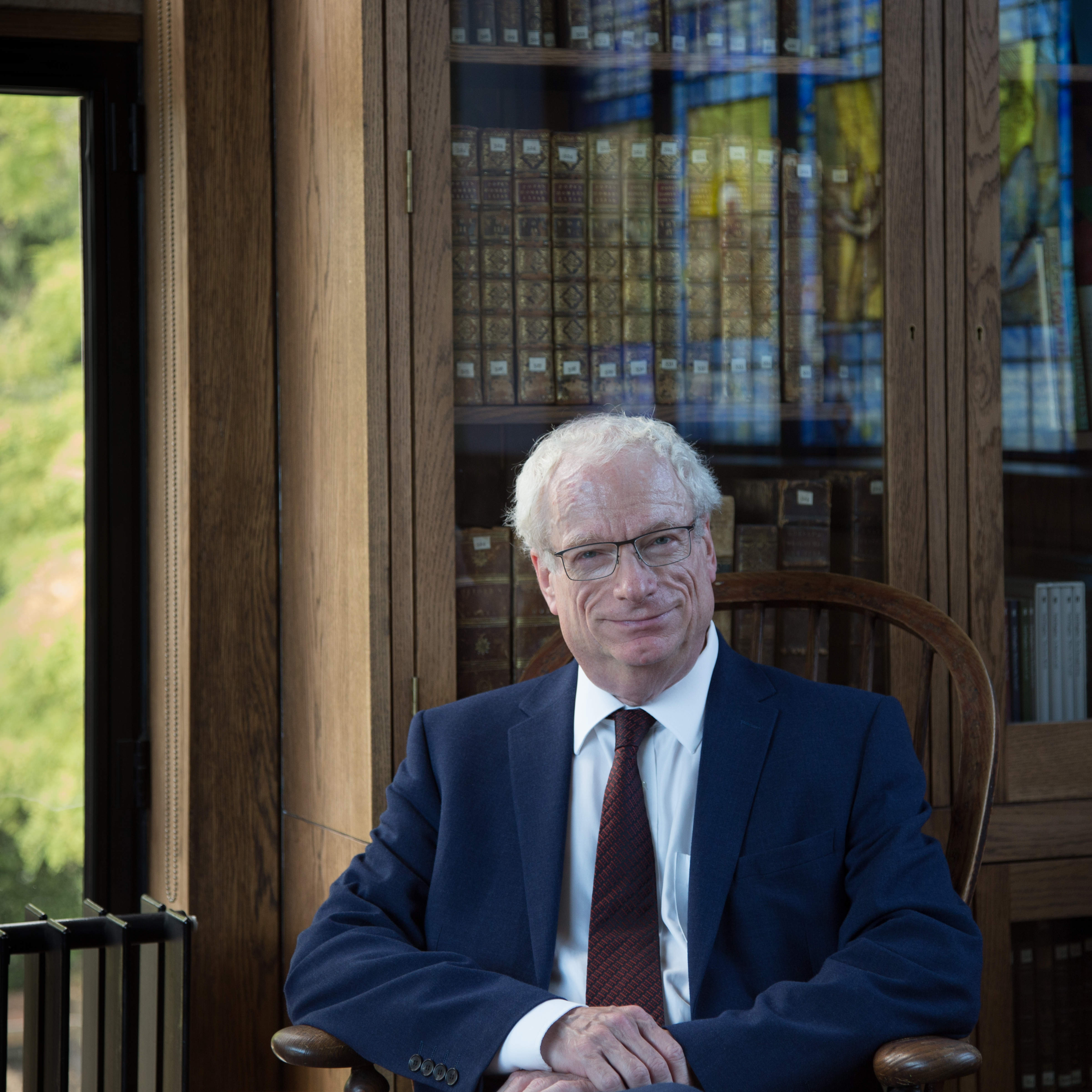 A photograph of an older white man with short white hair. He is sat in a chair wearing glasses and smiling at the camera. He is wearing a blue suit with a white shirt and red tie. His hands are resting on his lap. The background is a bookcase with glass doors.