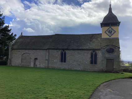 A photograph of a church building. It is one long rectangle with one tower at the end of the building. The tower has a clock on it. It is a plain building with plain wooden doors and plain glass windows.