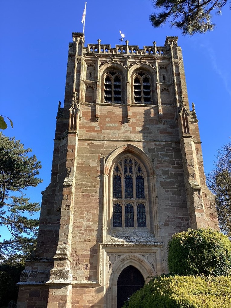 A photograph of a tower of a church with a flag on top. The sky behind is clear blue.