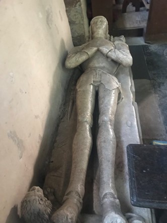 A photograph of the full body effigy of Sir Hugh Mortimer. He is laying down with an animal by his feet. He is wearing armour and has his hands folded on his chest.