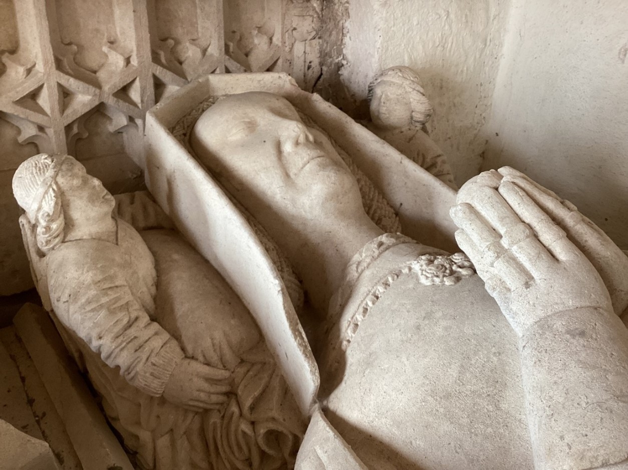 A photograph of the head of the effigy for Eleanor Croft. She has her hands together holding (possibly) a flower. She is wearing a headdress and necklace. There are two small figures looking on her by her head.