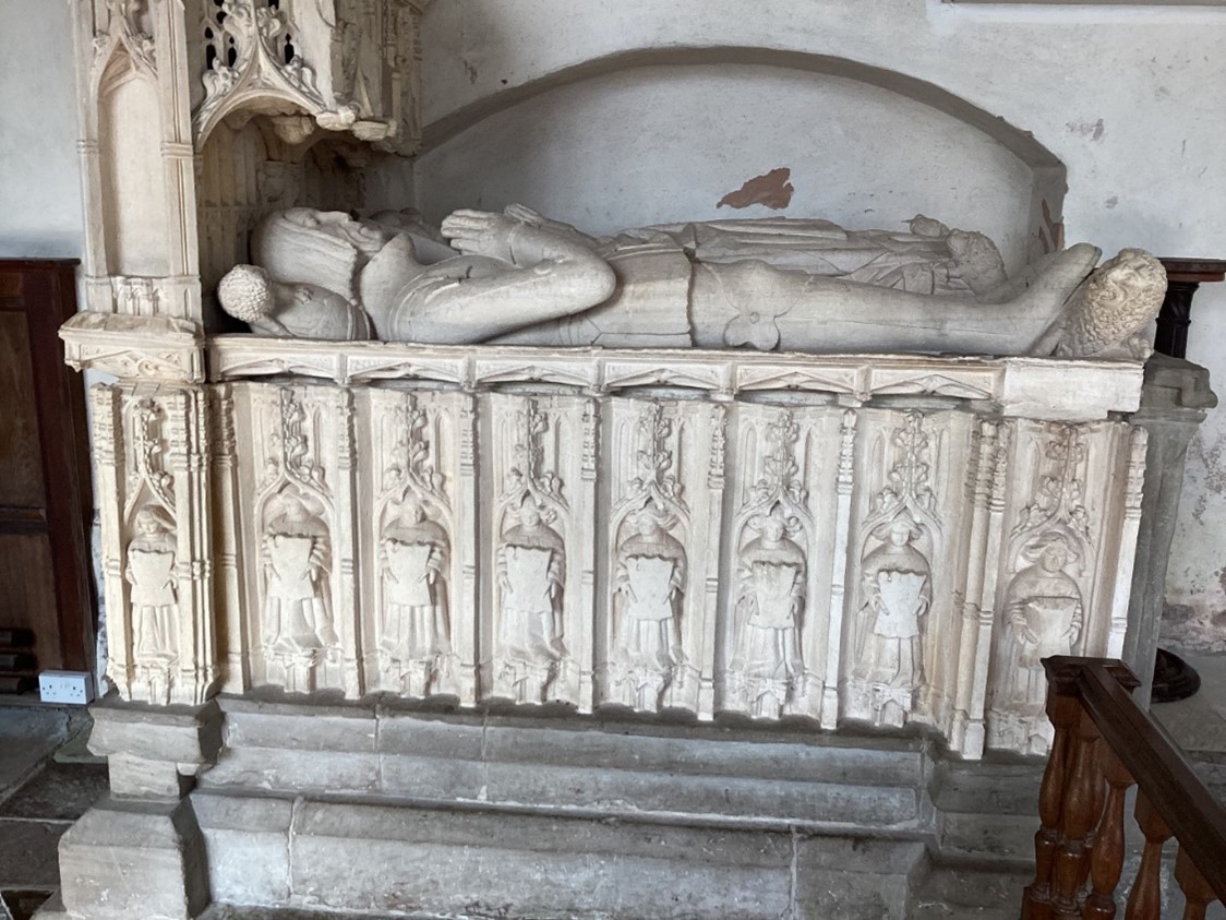 A photograph of an effigy of a person lying down with his hands together in prayer, there is a lion by his feet. He is laying on a tomb that has been carved.