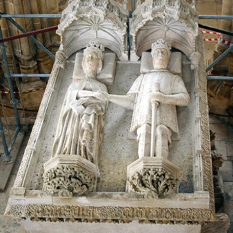 A photograph of an effigy of two people lying side by side. The women on the left has a crown and is lying on a pillow, the man on the right is wearing a crown, holding a sword, and is lying on a pillow. They are holding hands.