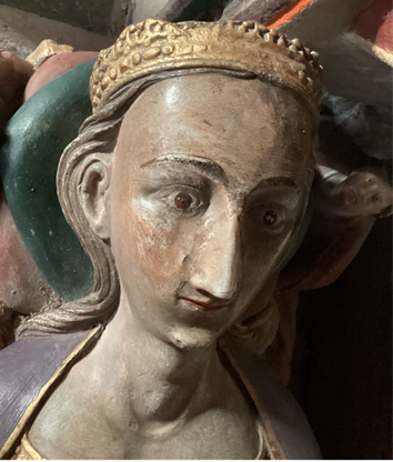 A photograph of the head of an effigy. She is wearing a crown, has long brown hair and is wearing purple. Her eyes are open.