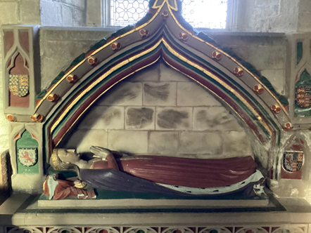 A photograph of an effigy. A woman is lying down with her hands together in prayer. She is wearing a read dress and a robe. 