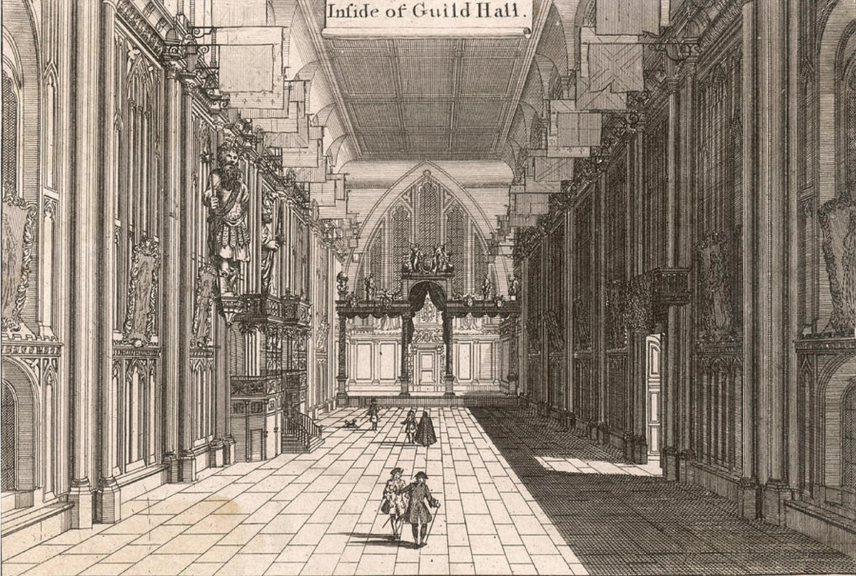 Engraving of the view inside London's Guildhall. Interior of a hall, with flags along the ceiling, and five figures walking towards the end of the room.