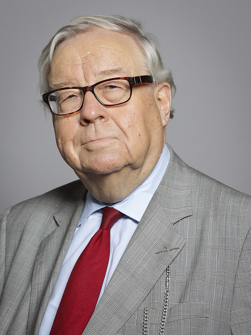 Photograph of Lord Cormack. He has grey hair and is wearing tortoiseshell glasses. He is wearing a grey checked suit jacket, a blue shirt and red tie. 