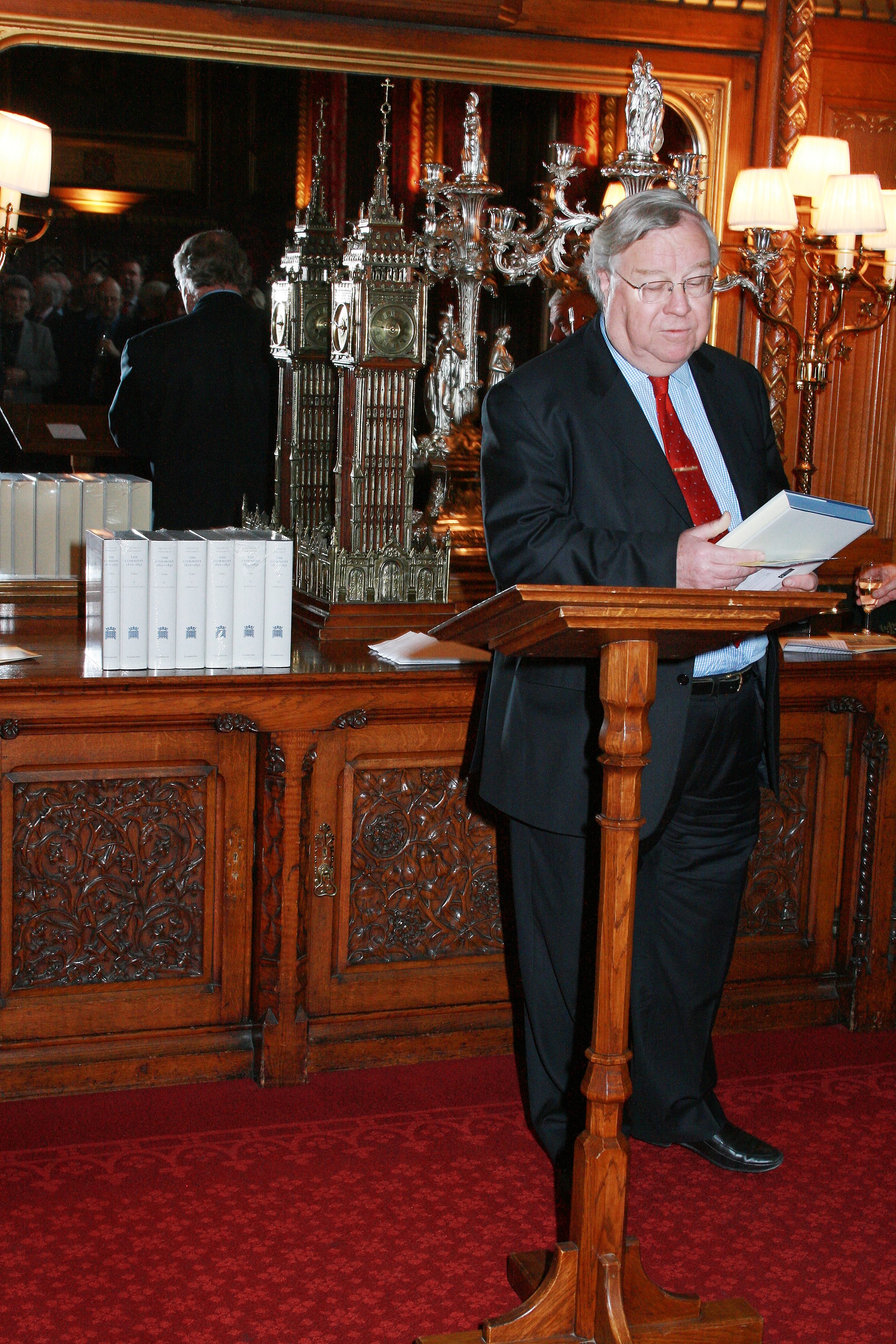 Photograph of Lord Cormack. He is standing in a dark wood panelled room with red carpet, in front of a wooden lectern. Behind him is a front of a large mirror and gold coloured scale model of the Elizabeth Tower (Big Ben) as well as a display of grey coloured books. Lord Cormack is wearing a dark suit, blue shirt and red tie. He is mid speech, holding one of the grey books (the House of commons 1820-1832) in his hands.