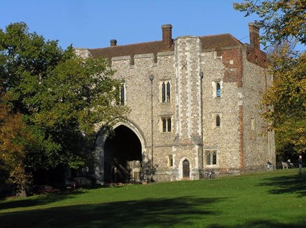 Photograph of a square stone building. It has a large arch in the centre, which acted as a gate. Slat windows are on the facade of the building and red brick chimneys are on the roof. The gatehouse is set on plush green grass with a tree to the left of the entrance.