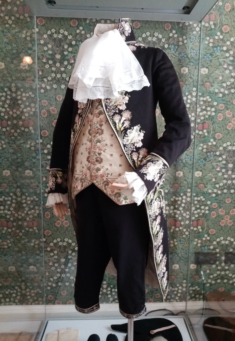 Picture of a restored suit. A torso mannequin is dressed with short navy trousers, a gold waistcoat, white shirt with large collar ruff and frilled cuffs, and an embroidered navy jacket. Floral embroidery is on the cuffs and edge of the jacket. The mannequin is in a glass display cabinet and there is floral wallpaper behind it.