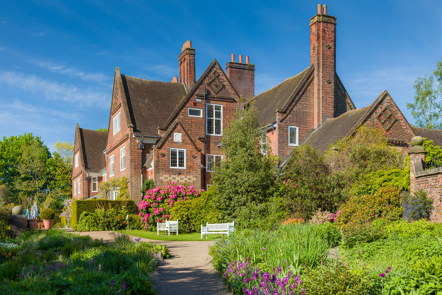 Photograph of Winterbourne House. A red brick building is surrounded by greenery and flowers. there are two benches in from of the house and a path winds around it.