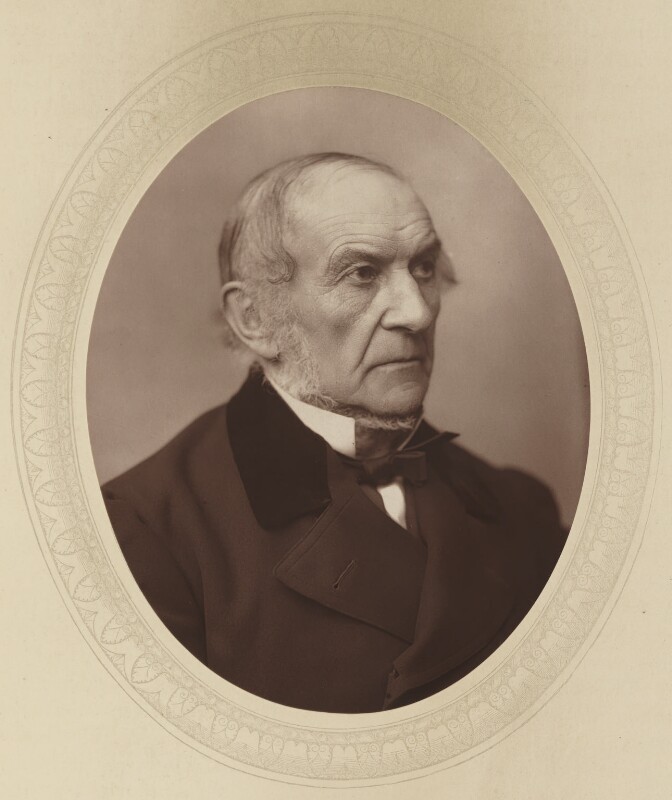An oval black and white picture of an older man, mounted on a cream backdrop with an egg and dart border around the photo. He is sitting side on, wearing a black suit jacket with a white shirt and a black bowtie. He has thinning hair and a grey beard on his sides and underneath his chin.