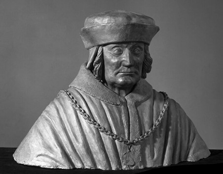 A photograph of a grey bust of a man from the chest up, shot against a slightly darker grey background. The man is wearing a robe with a chain across the front. He has long hair, just past his head, and is wearing a flat hat with a large rim folded upwards.