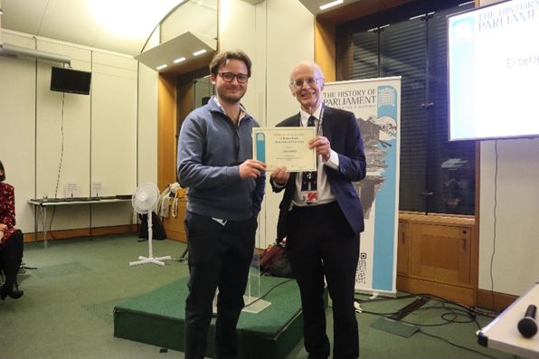 Two men are stood smiling at the camera, holding a certificate between them. Behind them is a banner with the words 'The History of Parliament' written on it.