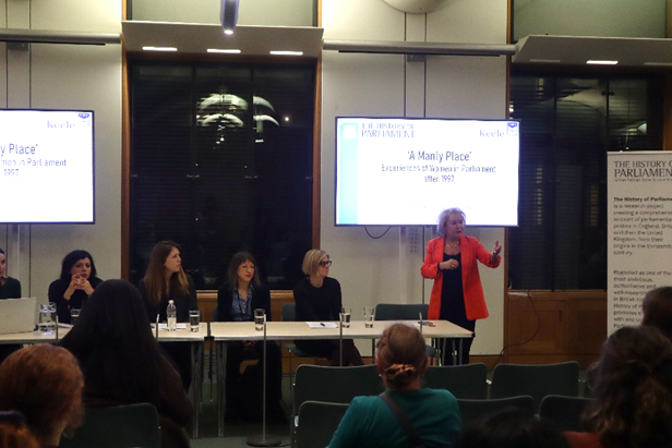 A panel of five women are sat behind a long desk, in front of a crowd of people. Another woman is stood up in the centre of the room, wearing a bright orange jacket. She is mid-speech and gesturing to the crowd who are looking at her. Behind the panel are two screens displaying the title of the event, 'A Manly Place: Experiences of Women in Parliament after 1997'.