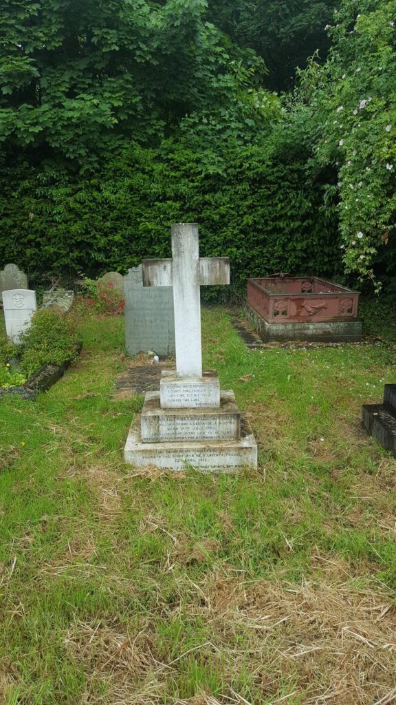 A picture of William Glynne Charles Gladstone's grave. In the middle of the picture stands the grave with a white cross on top, with a three tiered plinth with text on. It is surrounded by green grass and behind the grave is a darker green hedge. 