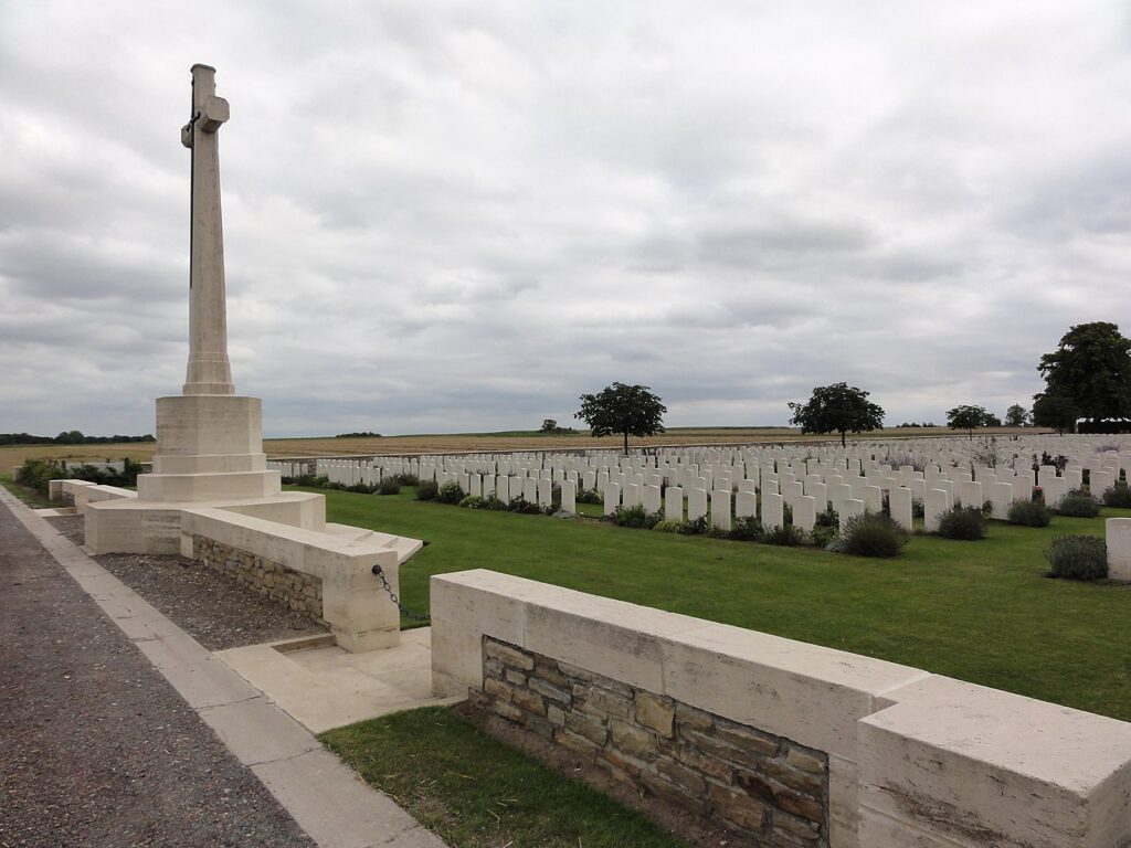A graveyard in a field, with a large cross at the front of the cemetery, overlooking a field full of white uniform gravestones. 