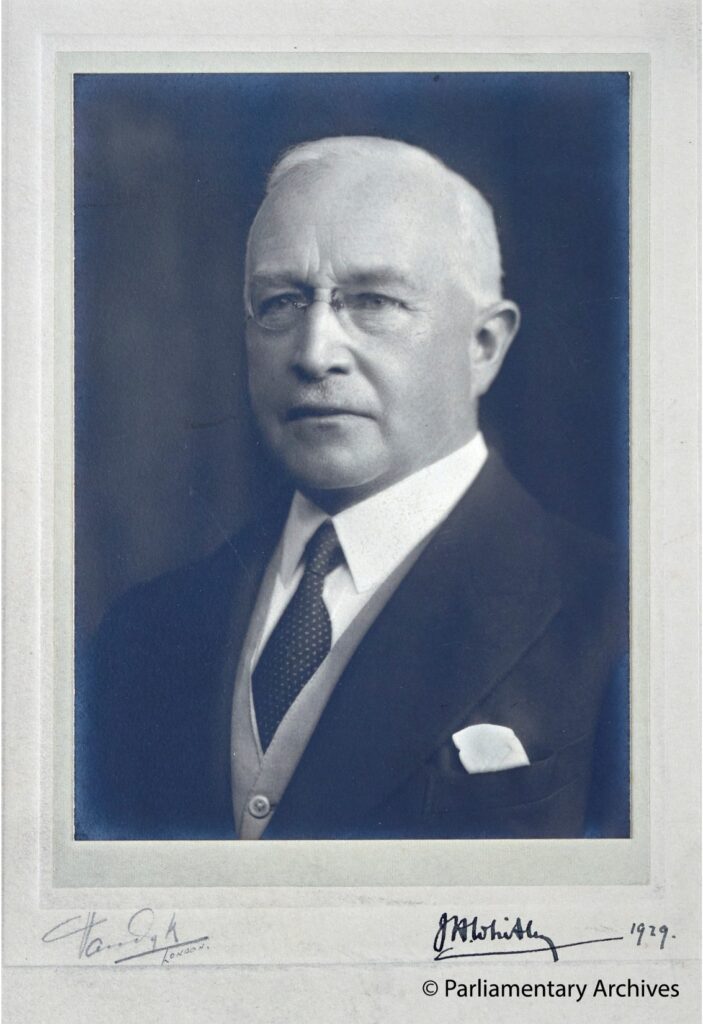 A head and shoulders profile of a man with white hair and spectacles in a suit.