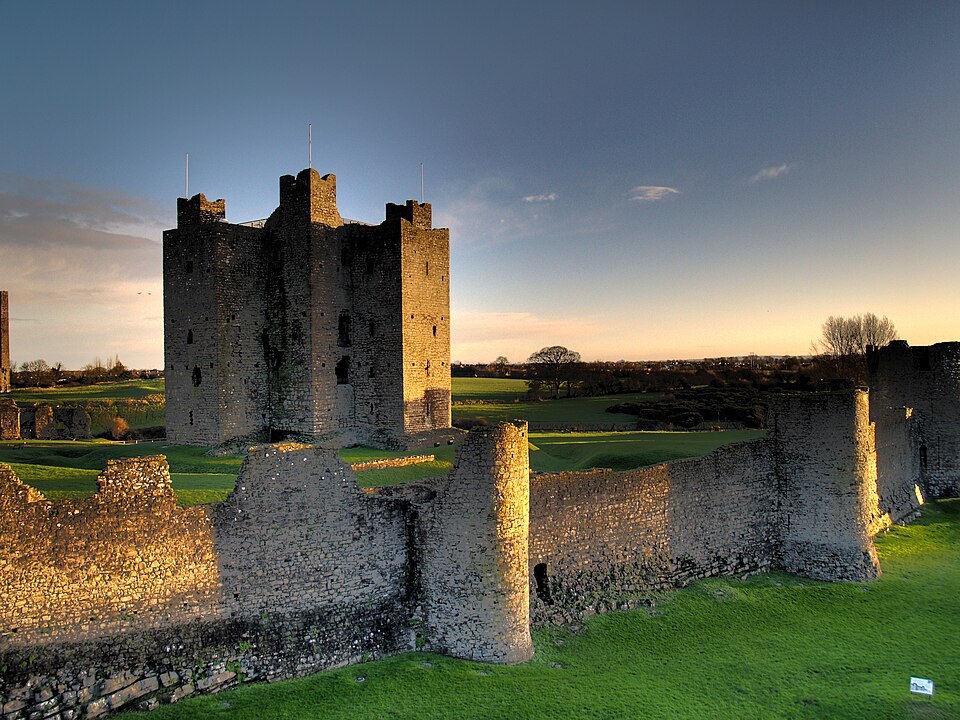 A coloured photograph of Trim Castle. Taken at sunrise, warm light covers the parts of the castle facing the sunrise. In a green setting away from any roads, the castle in the centre is not relatively tall and has three small and flat turrets. The castle is surrounded on the right and in the foreground of the image by a low remains of a wall with two circular parts which used to be turrets. 