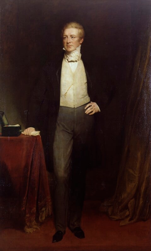 A full-lengthe portrait of Sir Robert Peel. Standing with his right hand resting on a red tableclothed table, He is wearing black shoes, dark grey trousers, a cream waistcoat, white shirt with a thick white necktie and high collar, and a black knee-length jacket. He is clean shaven with side parted short blonde hair. 