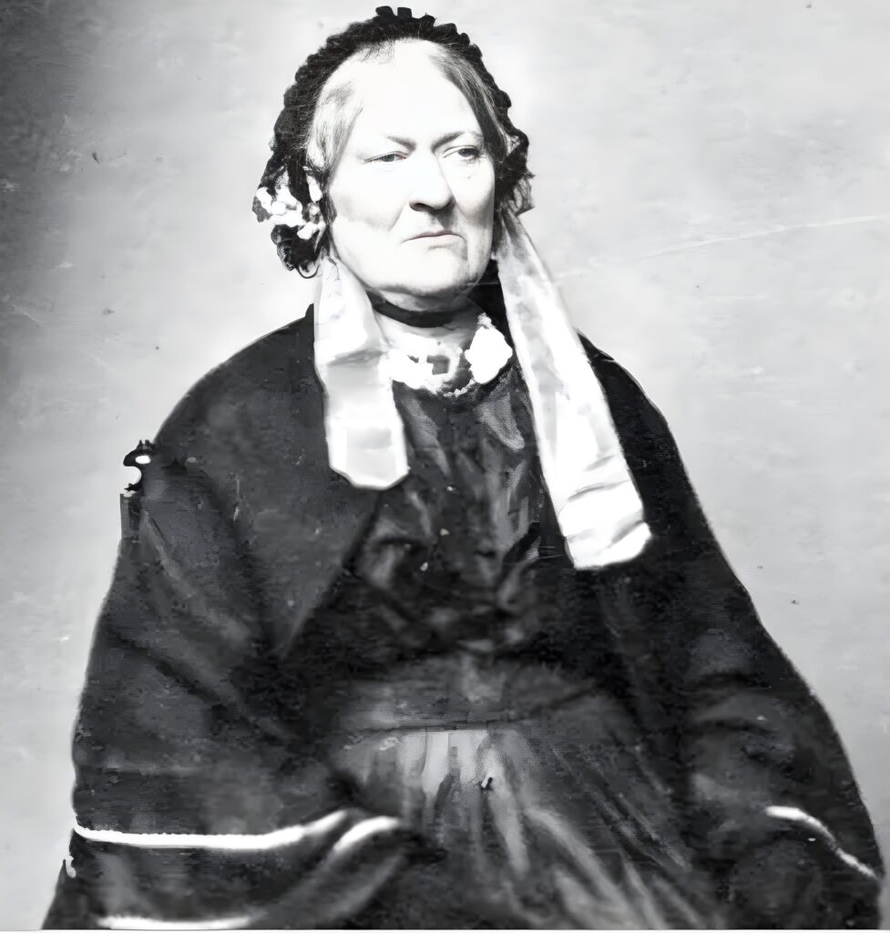 A half-length photographic black and white portrait of Lily Maxwell. Dressed in all black with a black skirt blouse, skirt, shawl and gloves, she is wearing a black bonnet with white material either side over her shoulders. She has grey hair.  