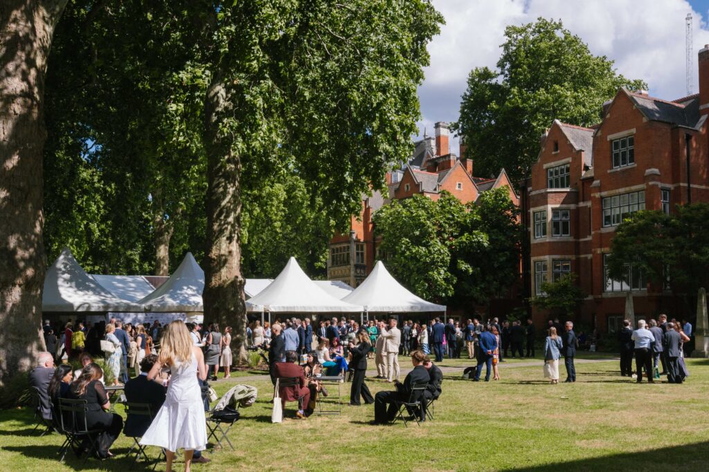 A photograph of the monarchy and democracy event. In a garden, with red bricked houses lining one side, bathed in sunlight, the garden is full of smartly dressed people, with a four peaked gazebo. To the left of the picture there are two tall and thick trees.