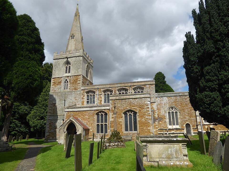 A photograph of St Peter's Church. It has a short sqyare tower to the left with a tall spire above the crenelated walls. To the right stretches the nave, with the church entrance next to the tower. In front of the church is a cemetery with green short grass. There are two tall dark green trees either side of the church. 